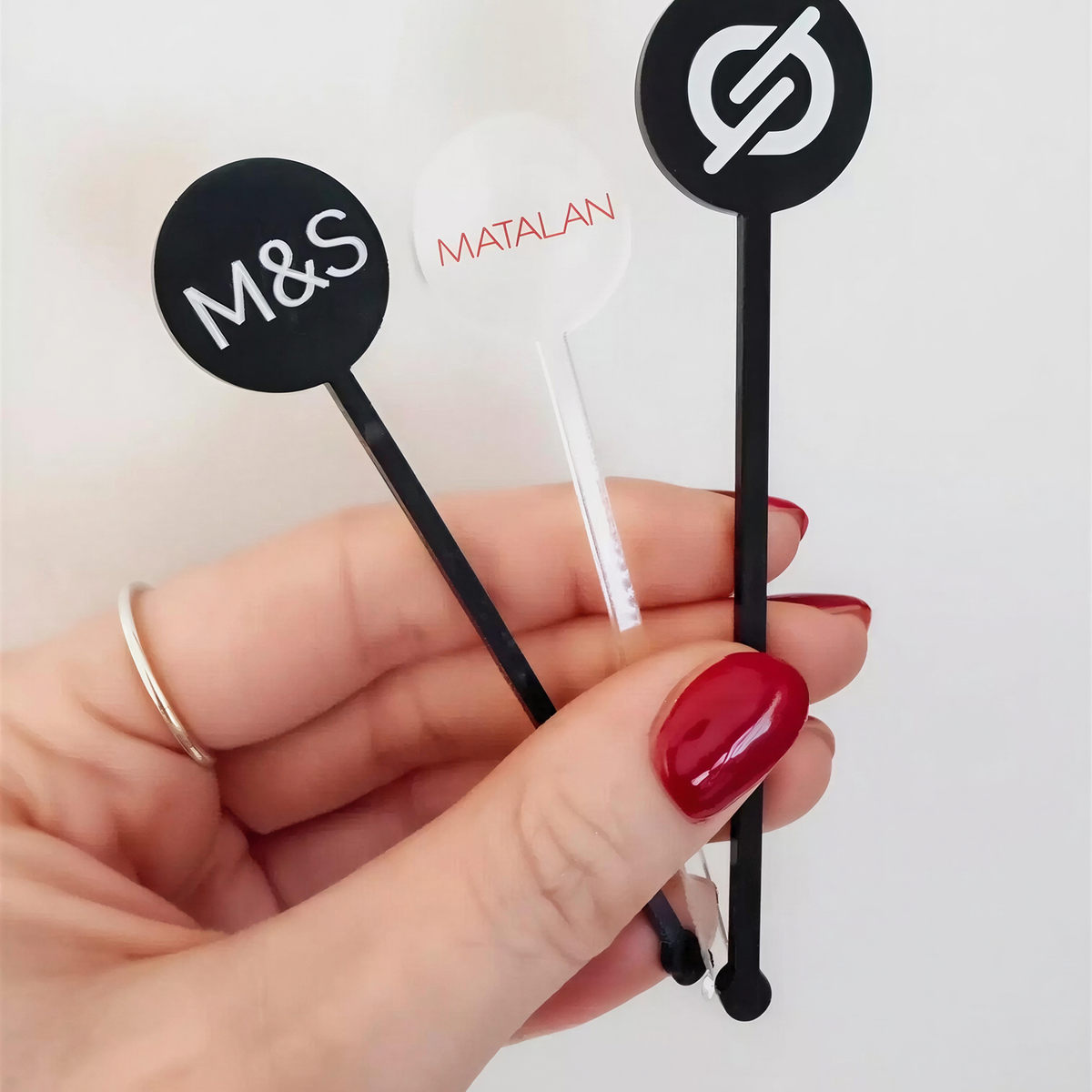 A hand holding three cocktail stirrers with different logos on them.