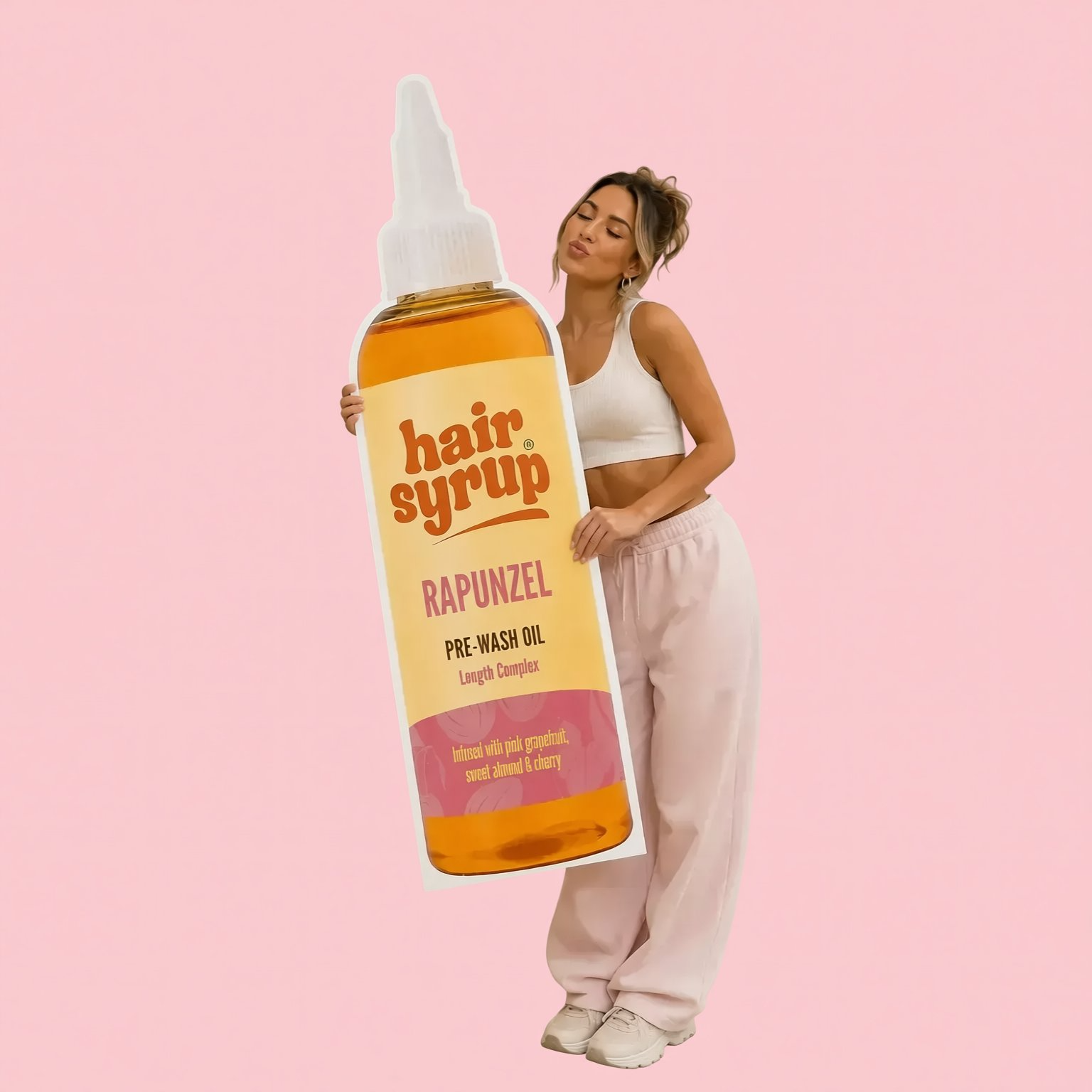 A woman is holding a large bottle of Rapunzel pre-wash oil in a room with a pink wall and a vase of flowers.
