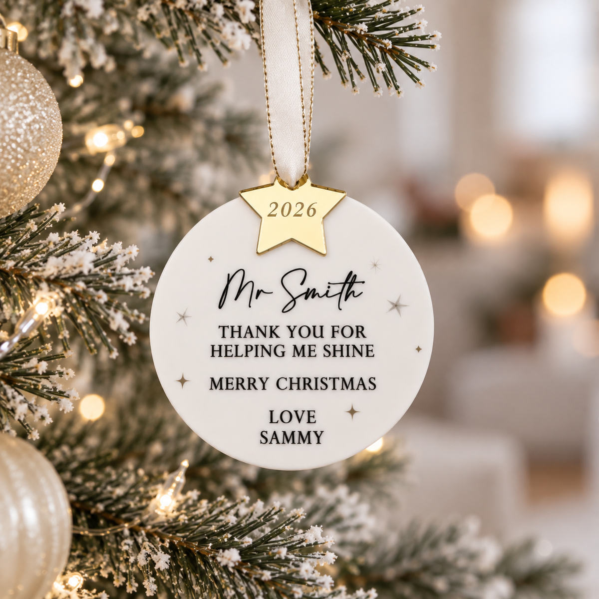 A white ornament with a gold star hanging from a Christmas tree, displaying a personalized message.