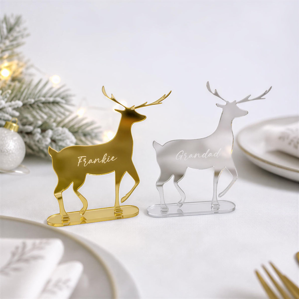 two decorative place holders in the shape of reindeer, one in gold and the other in silver, placed on a table with a white tablecloth and holiday decorations in the background.