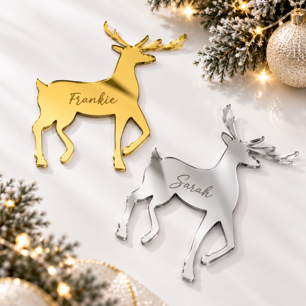 two personalized deer ornaments, one in gold and one in silver, placed on a white surface with Christmas lights and greenery in the background.