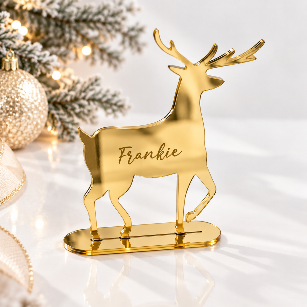 A golden deer-shaped ornament with the name "Frankie" engraved on it, placed on a white surface next to a Christmas tree with lights.
