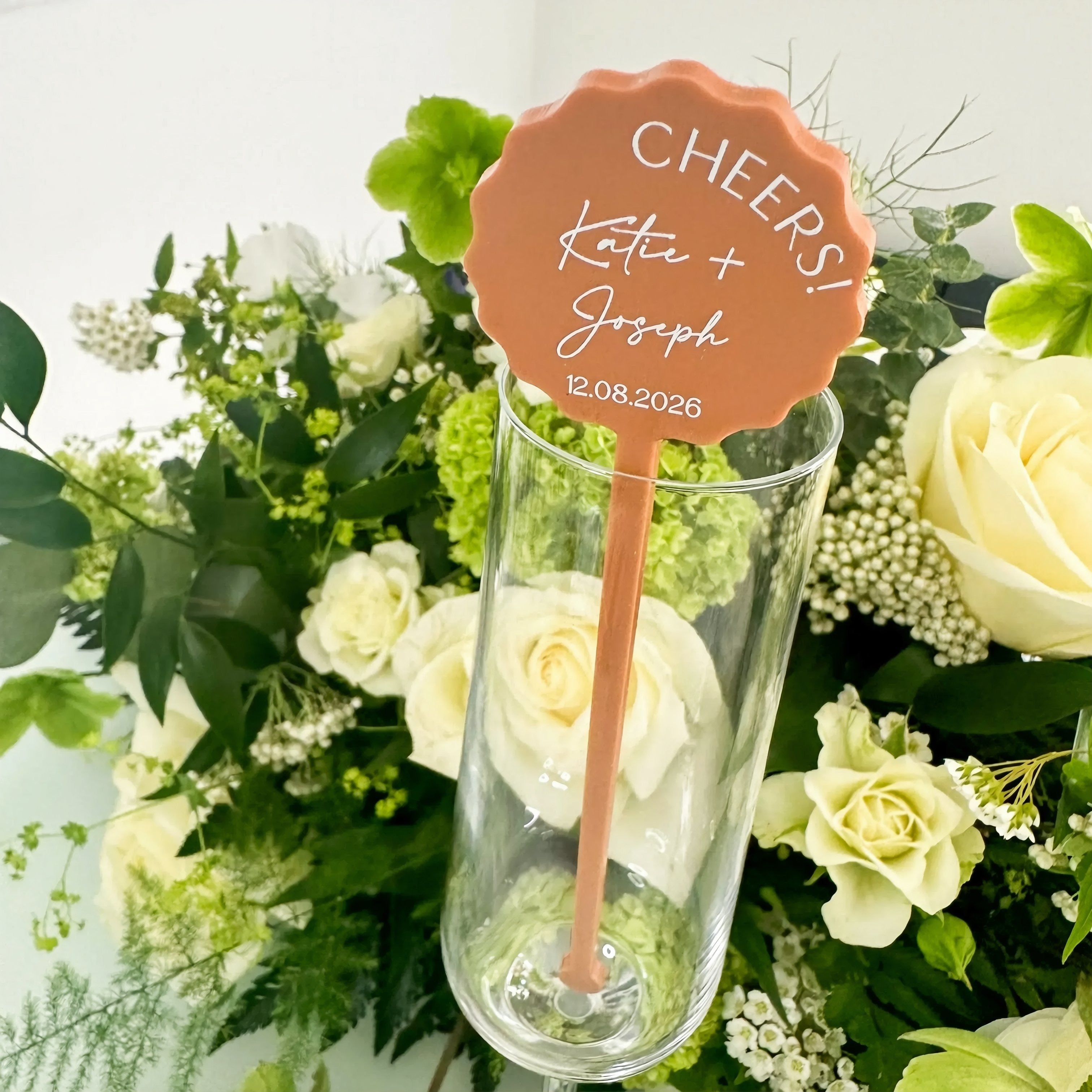 a glass with a decorative orange and white label attached to it, surrounded by a bouquet of white and green flowers.