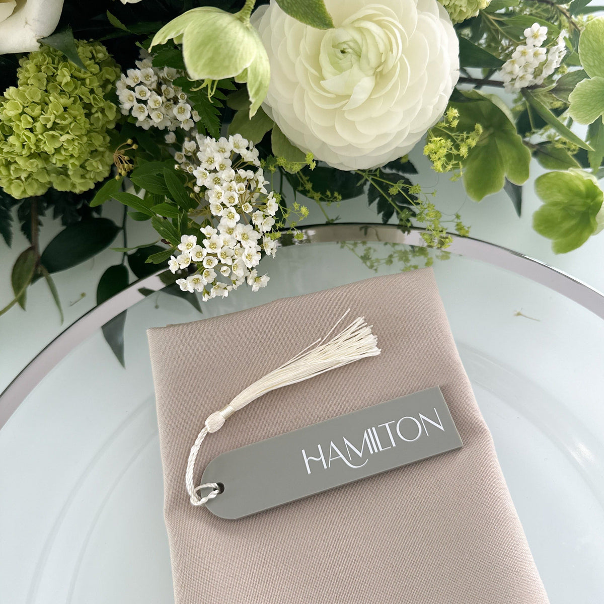 A collection of colorful name cards or place cards, each with a different color and text, placed on a white plate or napkin. The cards are arranged in a row, and there are flowers visible in the background.