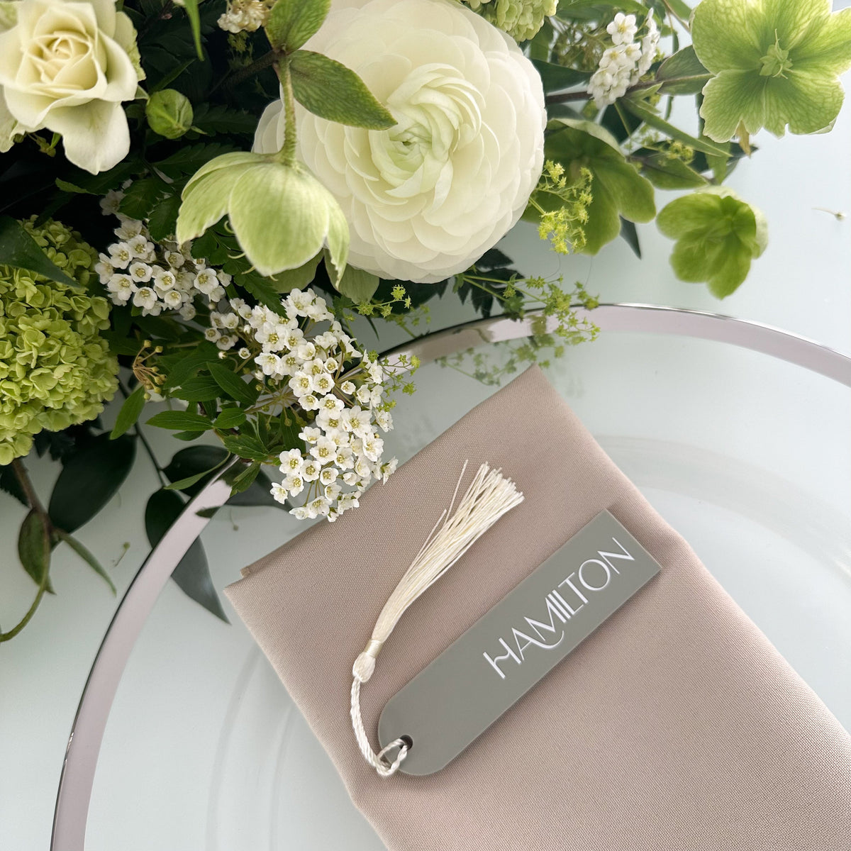 A gray name tag with the word "HAMILTON" printed on it, placed on a beige cloth with a white tassel.