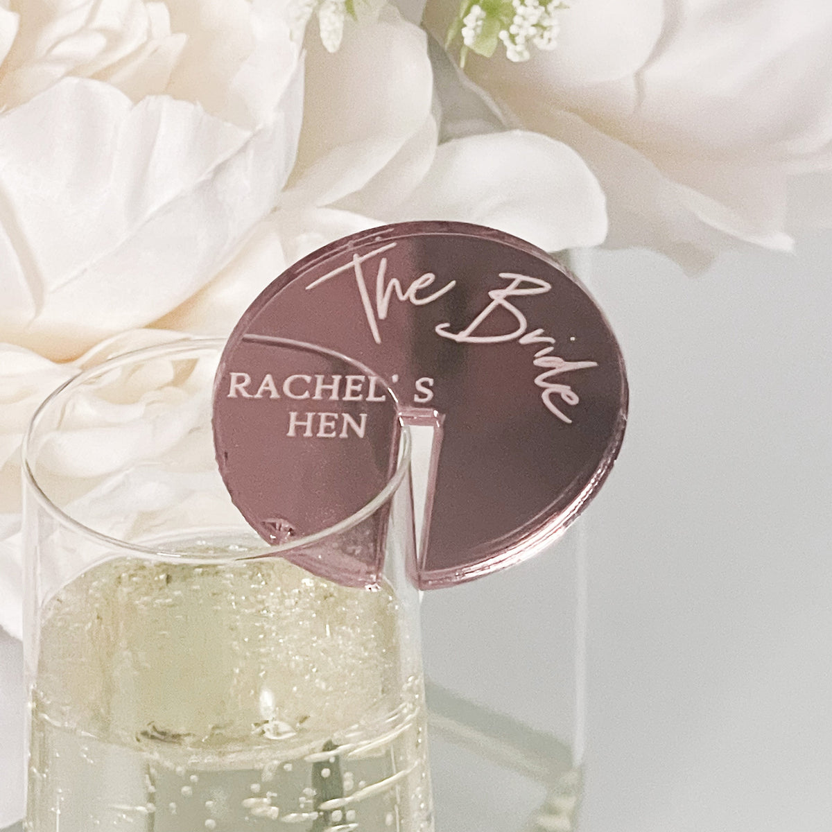 A glass of wine with a round coaster featuring the text "Mother of the Bride" and "RACHEL'S HEN" engraved on it, placed on a table with white flowers in the background.