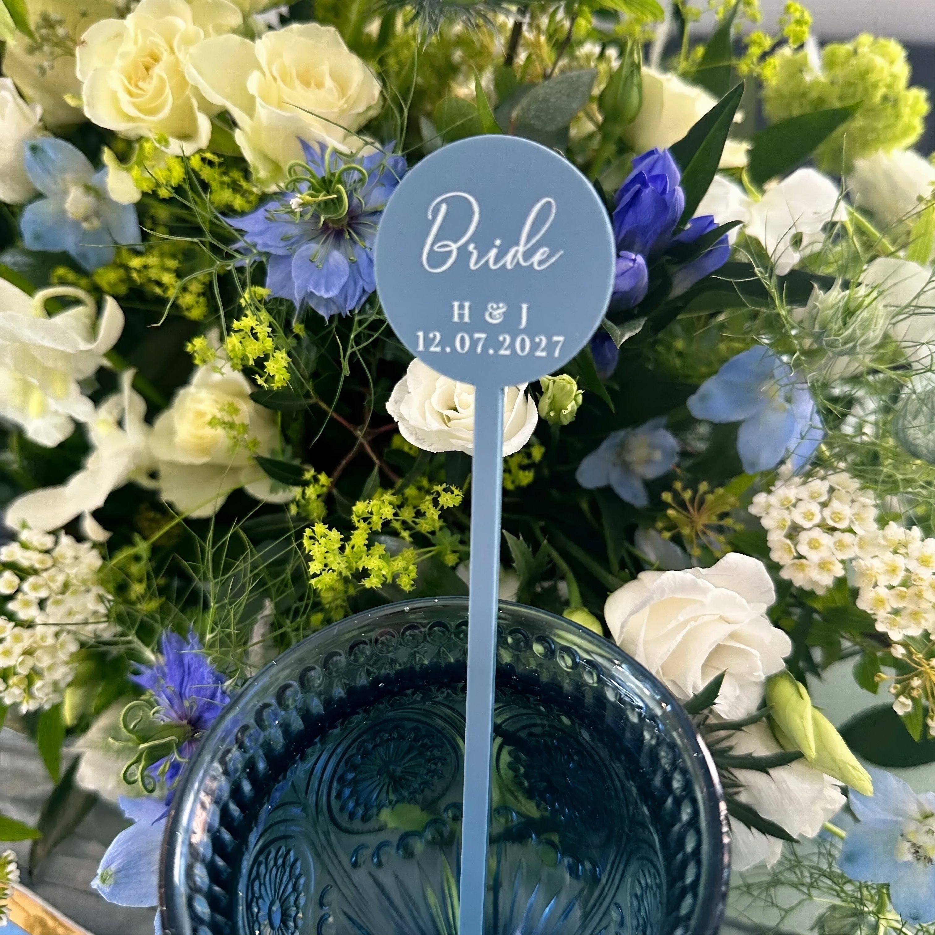 A blue wedding bouquet with white and blue flowers, and a blue circular sign with the text "Bride H & J 12.07.2027" placed in the center.