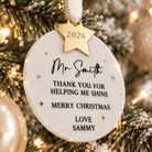 A white ornament with a gold star and the text "Mr. Smith" is hanging on a Christmas tree.