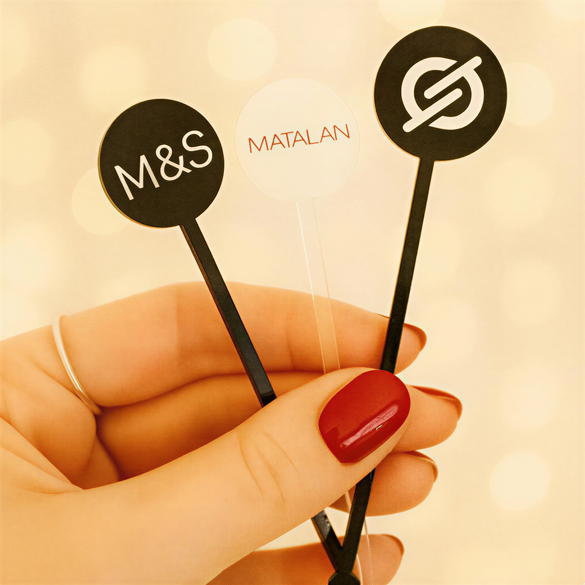 A hand holding three cocktail stirrers with the letters 'M&S' and 'MATALAN' printed on them.
