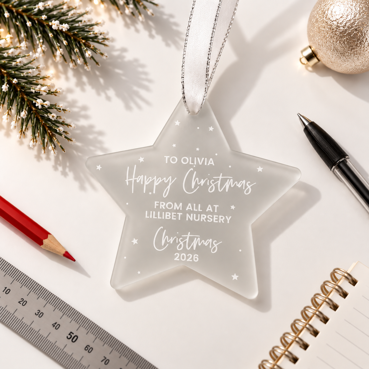 a clear star-shaped ornament with a personalized message, placed on a white surface alongside a ruler, a pen, and a Christmas ornament.