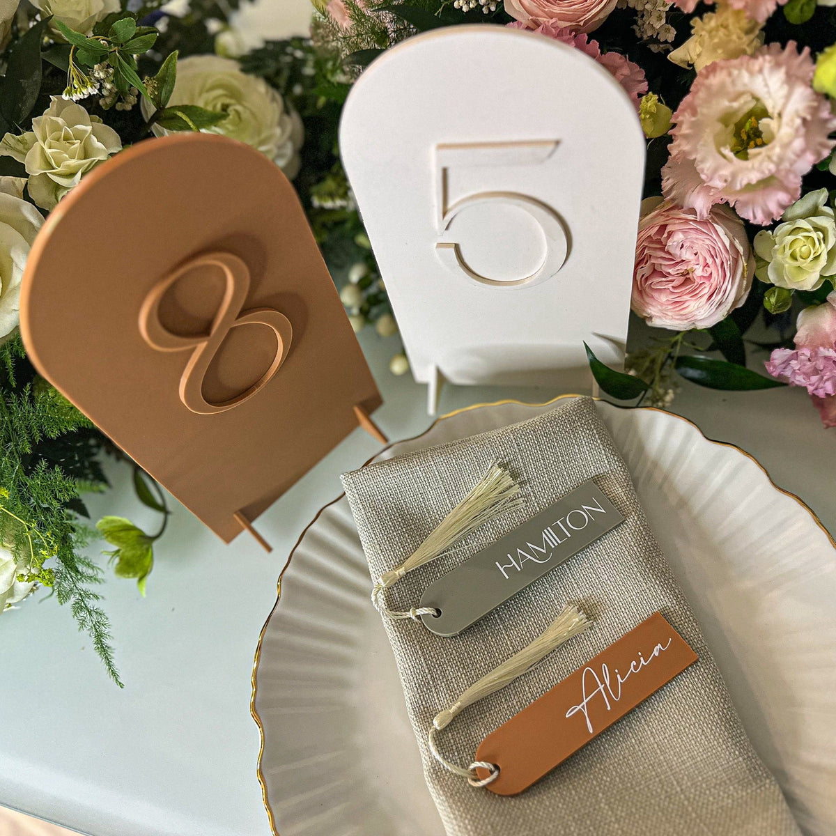 A wooden bookmark with the personalised name "Alicia" written on it, placed on a table next to a bouquet of pink and white flowers.