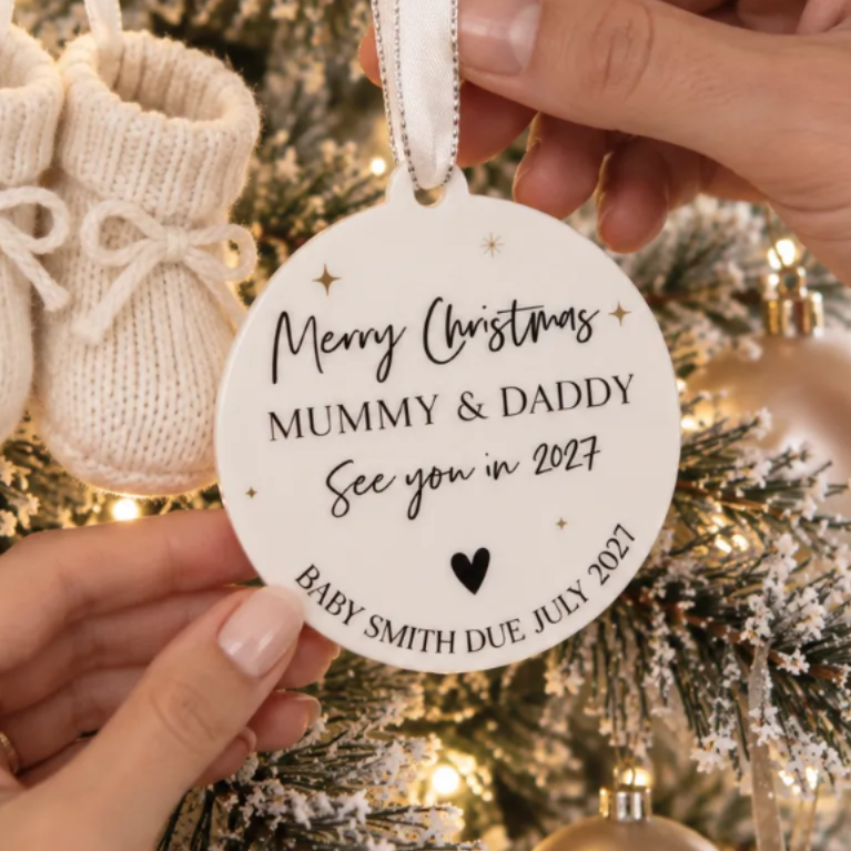 A person's hands are holding a white ornament with a message that reads "Merry Christmas MUMMY & DADDY See you in July 2021" on a Christmas tree decorated with white knitted booties and ornaments.