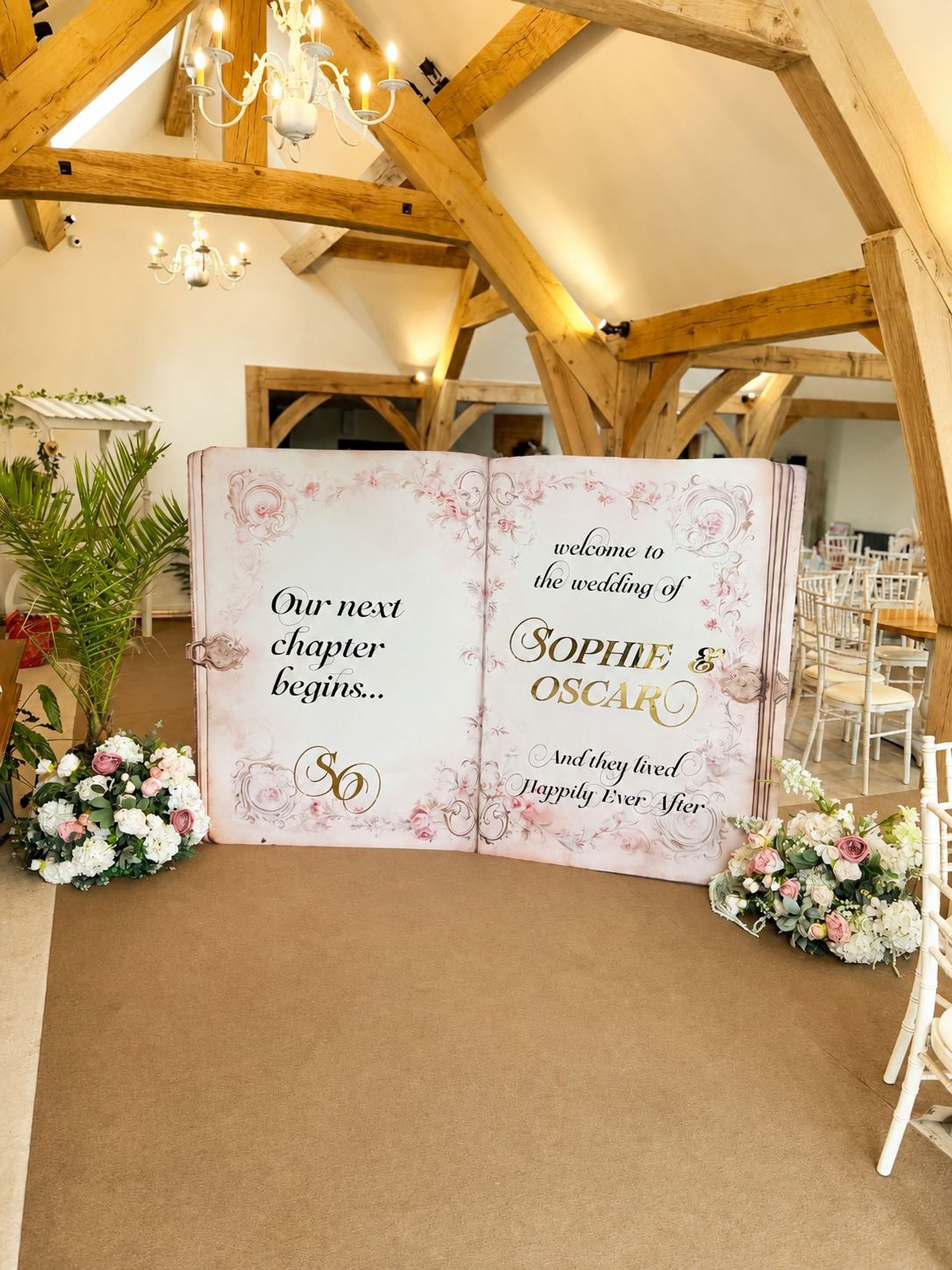 a large open book with a wedding invitation on the cover, surrounded by flowers and a wooden structure in the background.