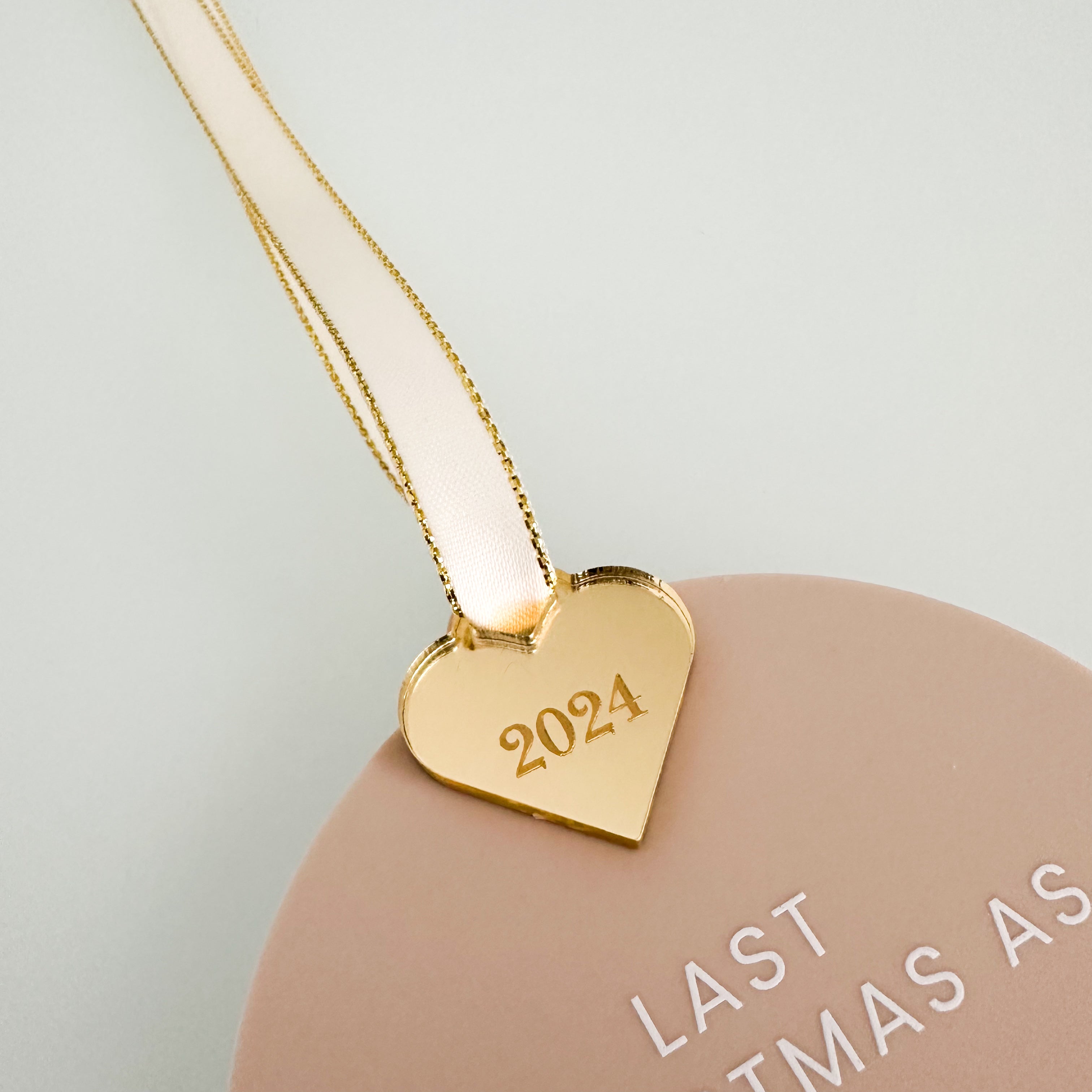 A white ornament with the text "LAST CHRISTMAS AS Miss Smith" and a heart-shaped ornament with the year "2024" on it, hanging on a Christmas tree.