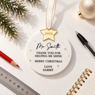 a white ornament with a gold star and a personalized message, surrounded by various office supplies such as a ruler, a pen, and a notebook.