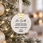 A white ornament with a gold star hanging from a Christmas tree, displaying a personalized message.