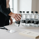 A person is holding a bottle of water from the "Interior Club" brand, with several identical bottles arranged on a table in front of them.