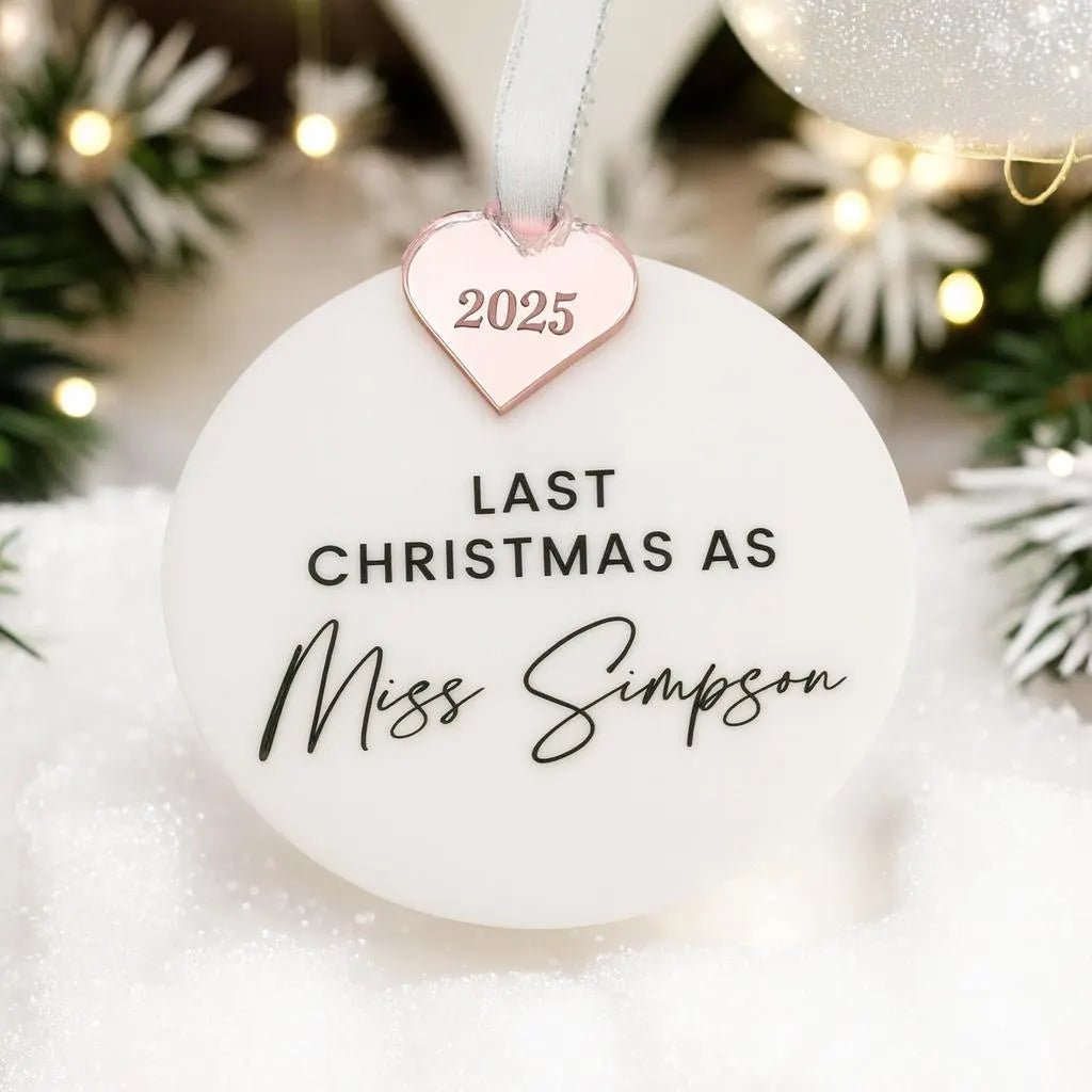 A round ornament with the text "LAST CHRISTMAS AS" with personalised "Miss name" and a heart-shaped charm with a year on it, hanging against a blurred background with Christmas lights.