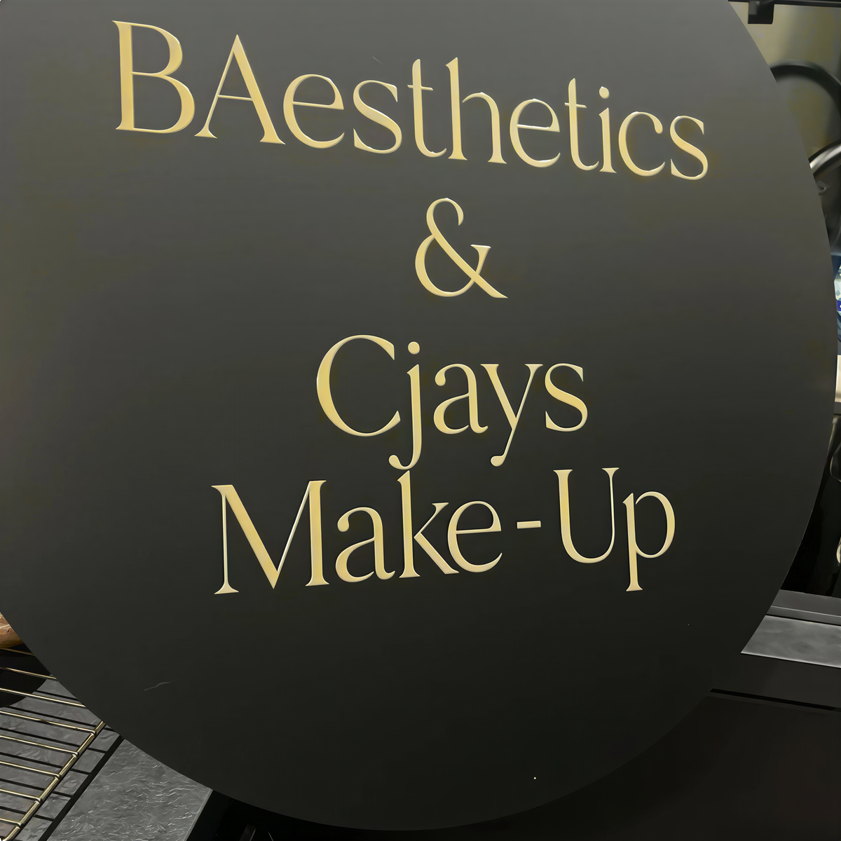 a black sign with gold lettering that reads "BAESTHETICS & CJAYS MAKE-UP".