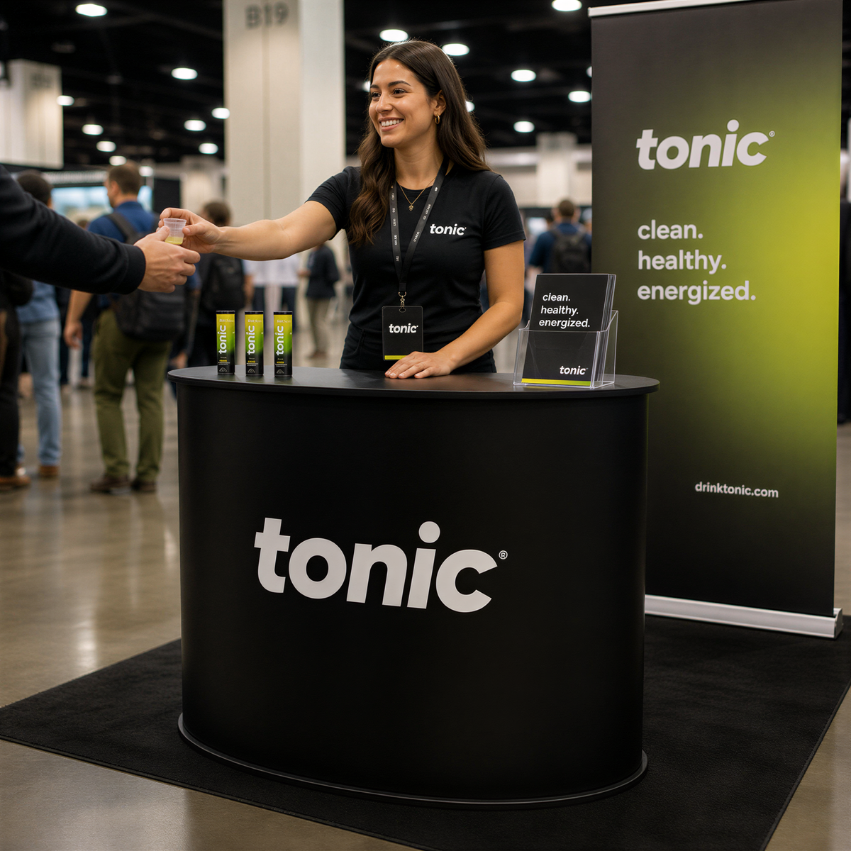A woman in a black Tonic-branded shirt is standing behind a black promotional booth, handing out samples of a product to a person.