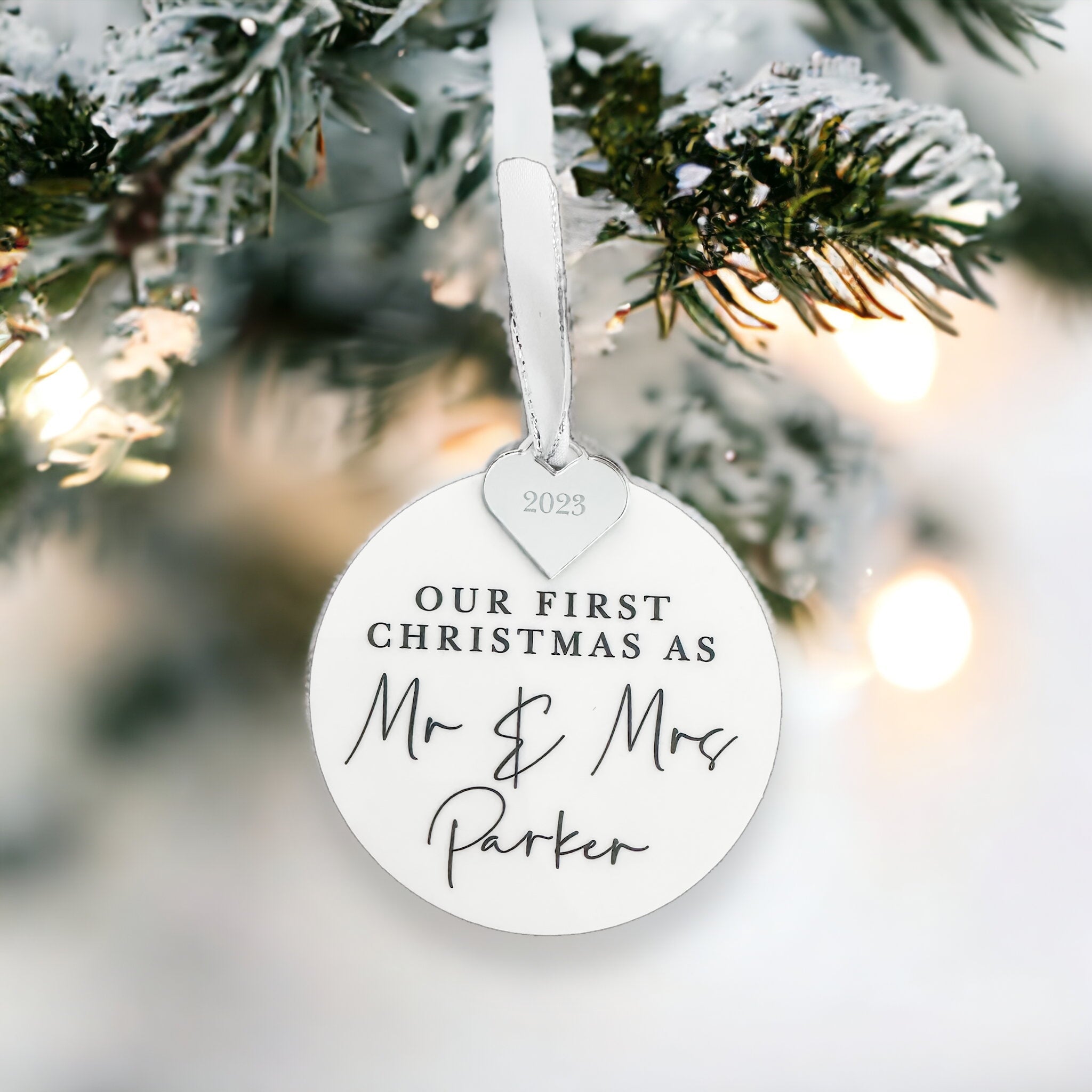 A black ornament with a gold heart-shaped charm hanging from a chain, displaying the personalised text "Our First Christmas As" and "Mr & Mrs Adams" written on it.
