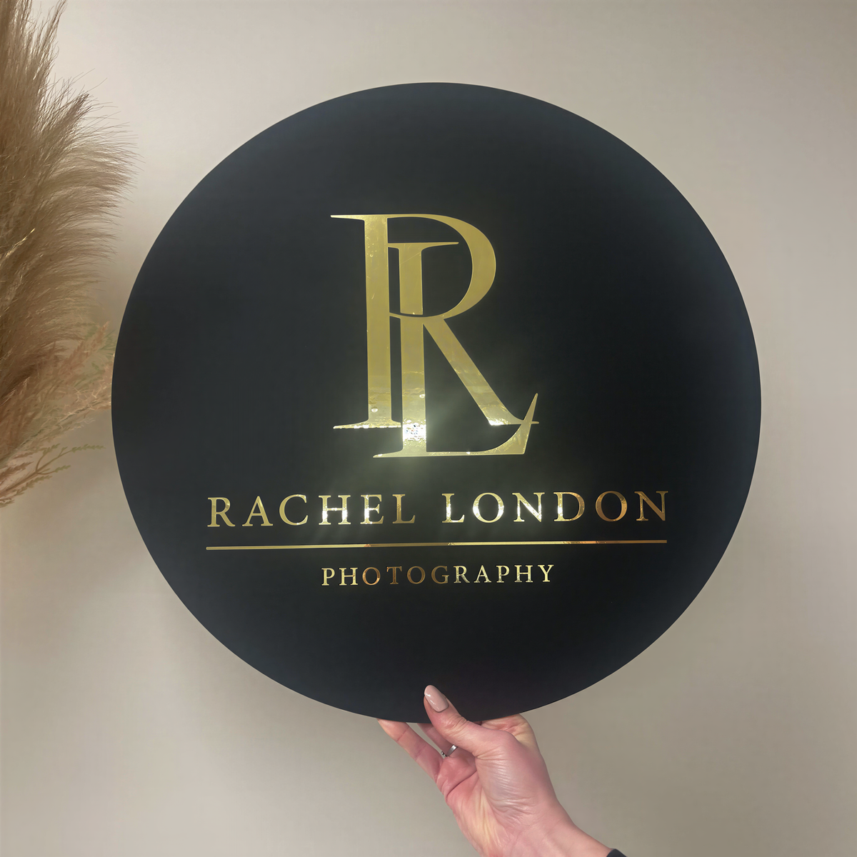 A person is holding a large black circular sign with the logo of Rachel London Photography.