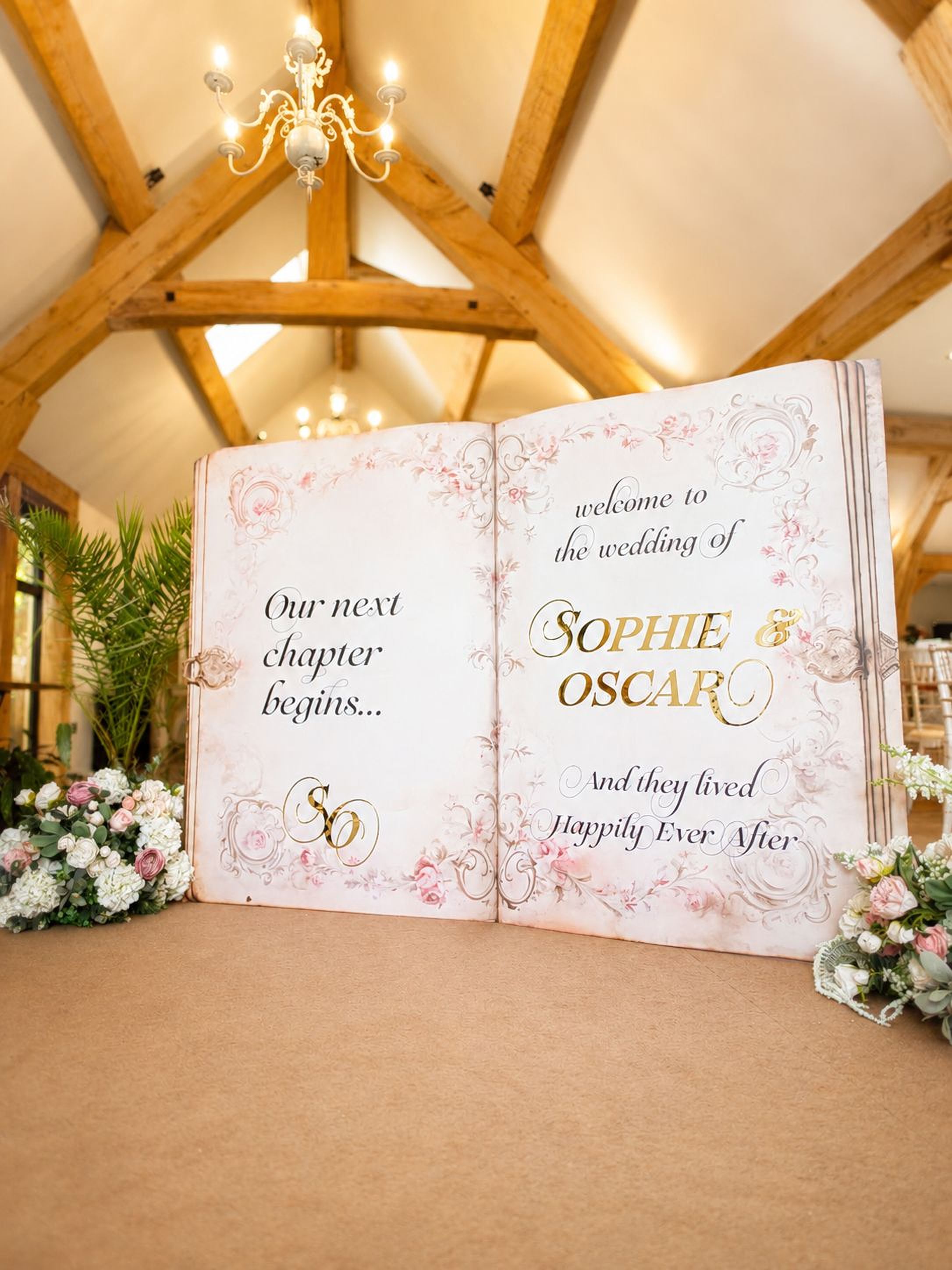 a large open book with a wedding invitation displayed on its pages, set against a wooden background with a chandelier overhead.