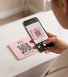 A person is holding a smartphone displaying a QR code on the screen, with a pink card featuring the same QR code in the background.