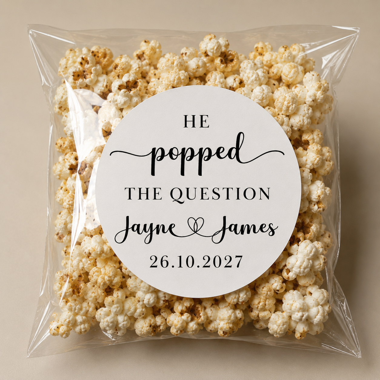 A clear plastic bag filled with popcorn, with a white circular label on it that reads "HE POPPED THE QUESTION Jayne & James 26.10.2027".