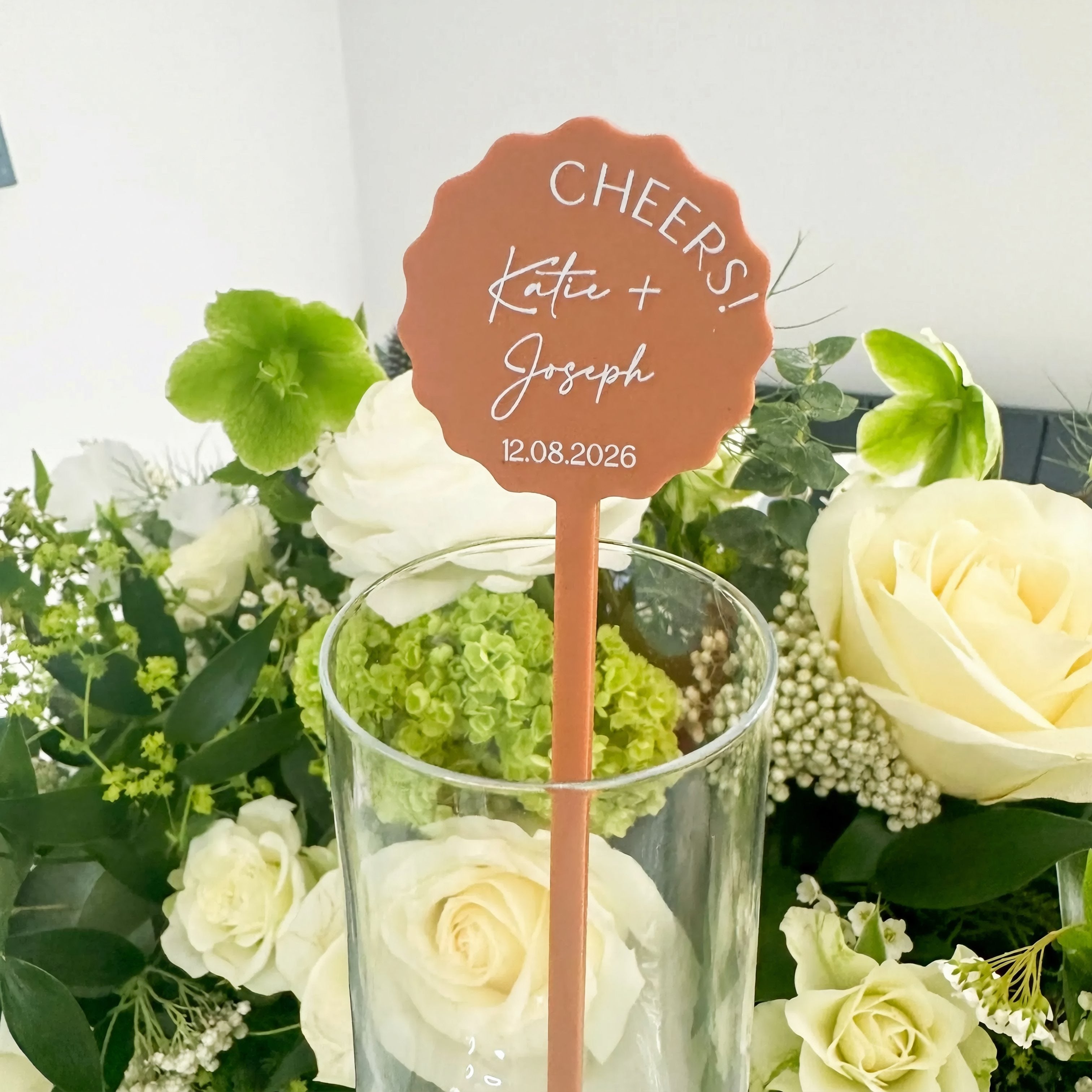 A hand holding a small orange card with the text "CHEERS! Katie + Joseph 12.08.2025" against a background of various colorful flowers.