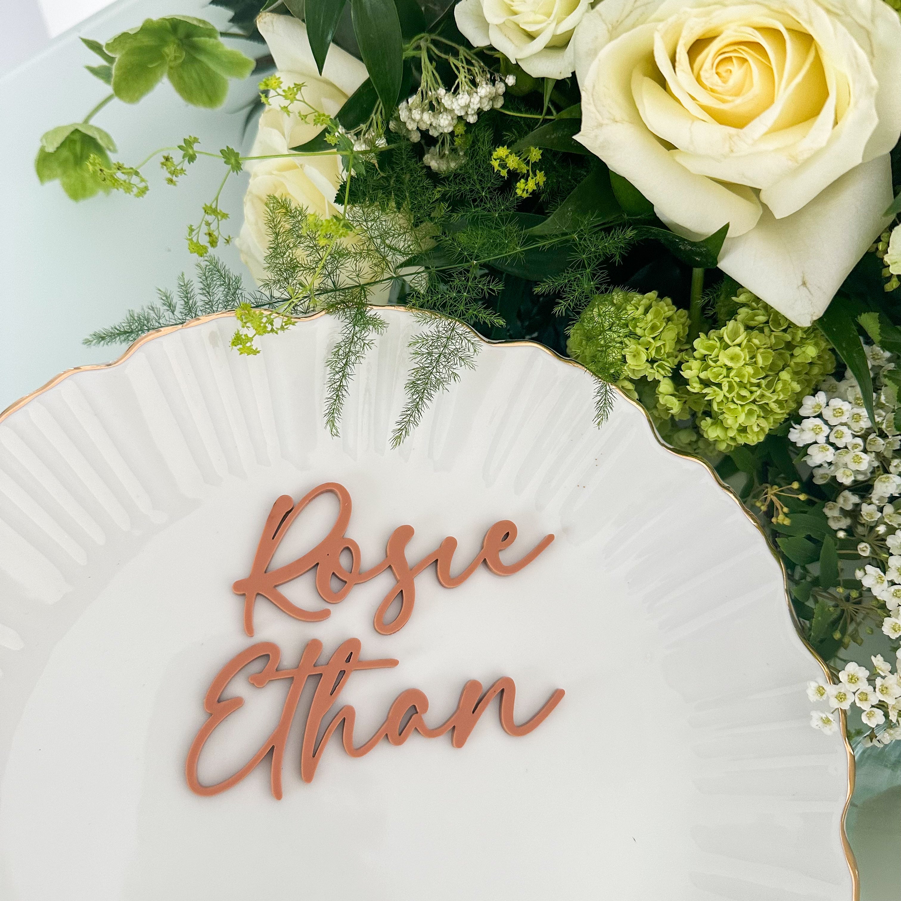 A collection of colorful name cards or place cards, each with a different color and text, placed on a white plate or napkin. The cards are arranged in a row, and there are flowers visible in the background.