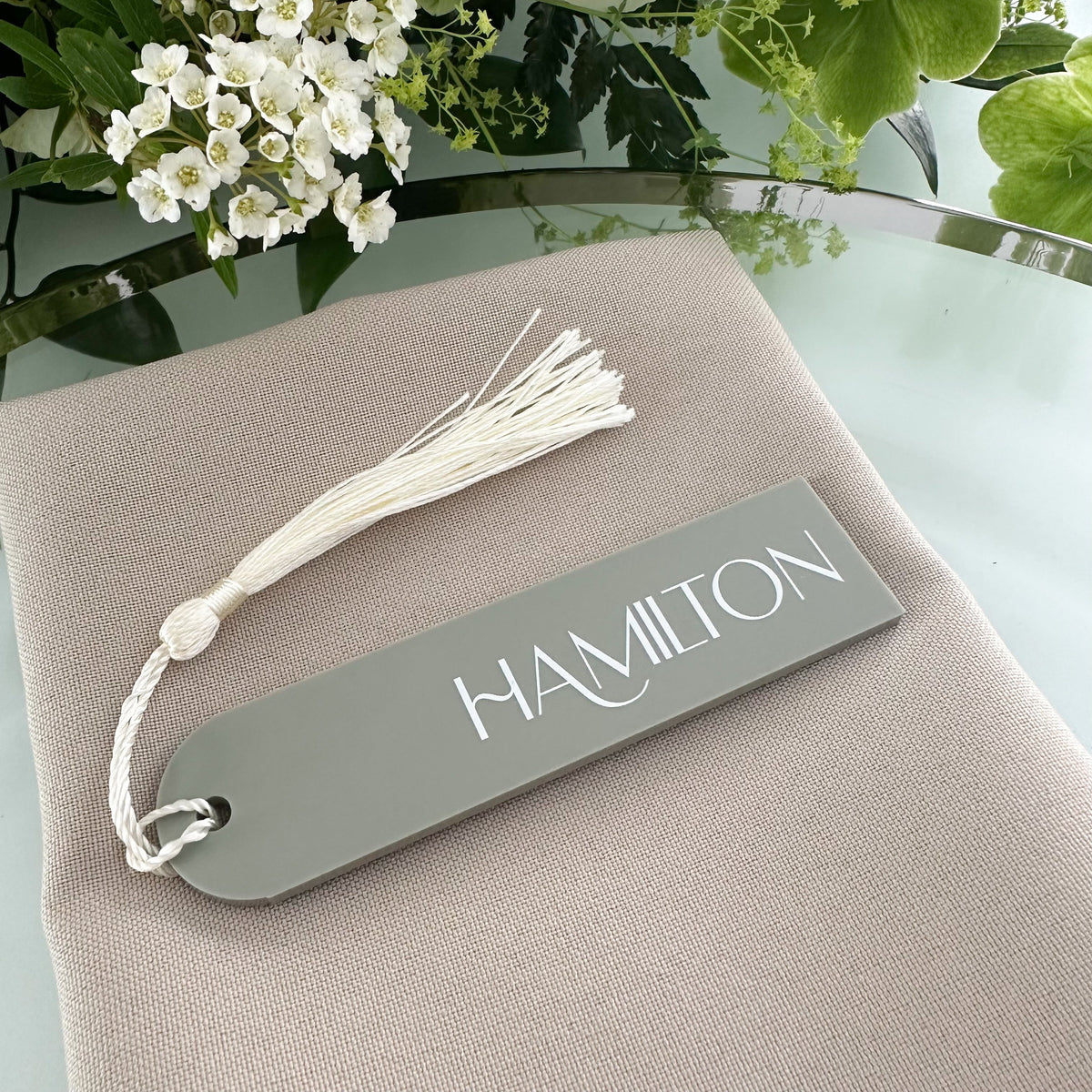 A table setting with a gray place card displaying the name 'HAMILTON' and a white tassel, surrounded by a floral arrangement of white and green flowers.