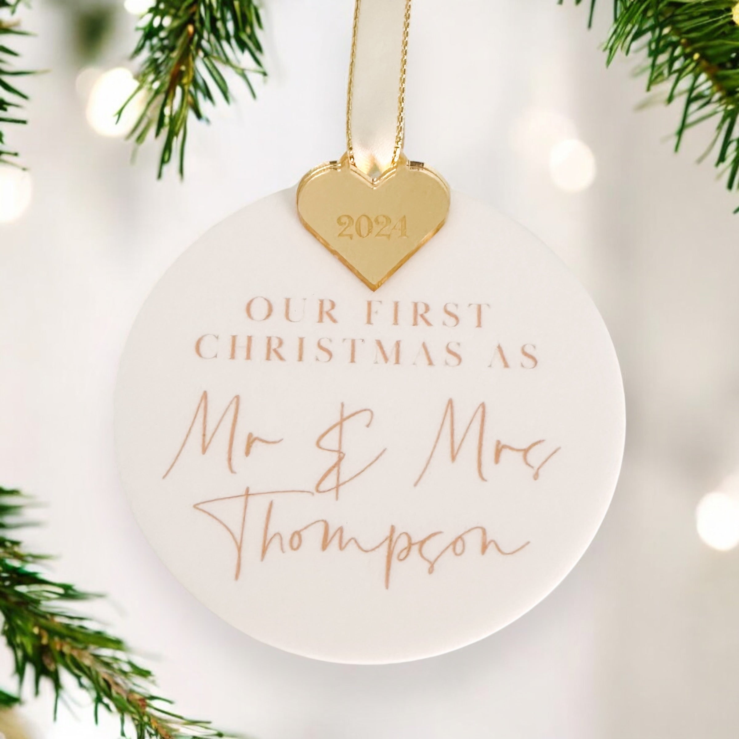 A black ornament with a gold heart-shaped charm hanging from a chain, displaying the personalised text "Our First Christmas As" and "Mr & Mrs Adams" written on it.