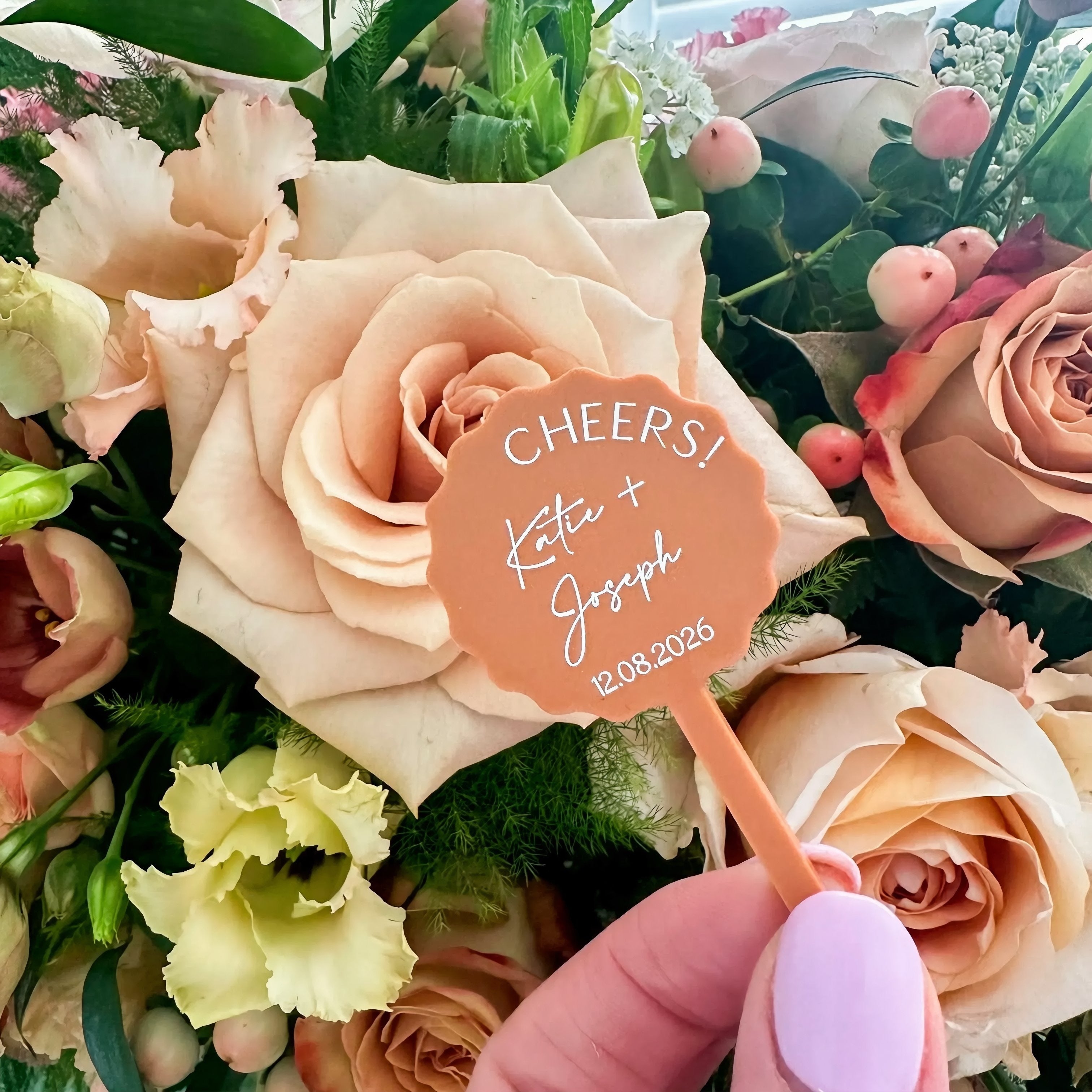 A hand holding a small orange sign with the text "CHEERS! KATE + JOSEPH" and the date "12.08.2026" against a background of various colorful flowers.