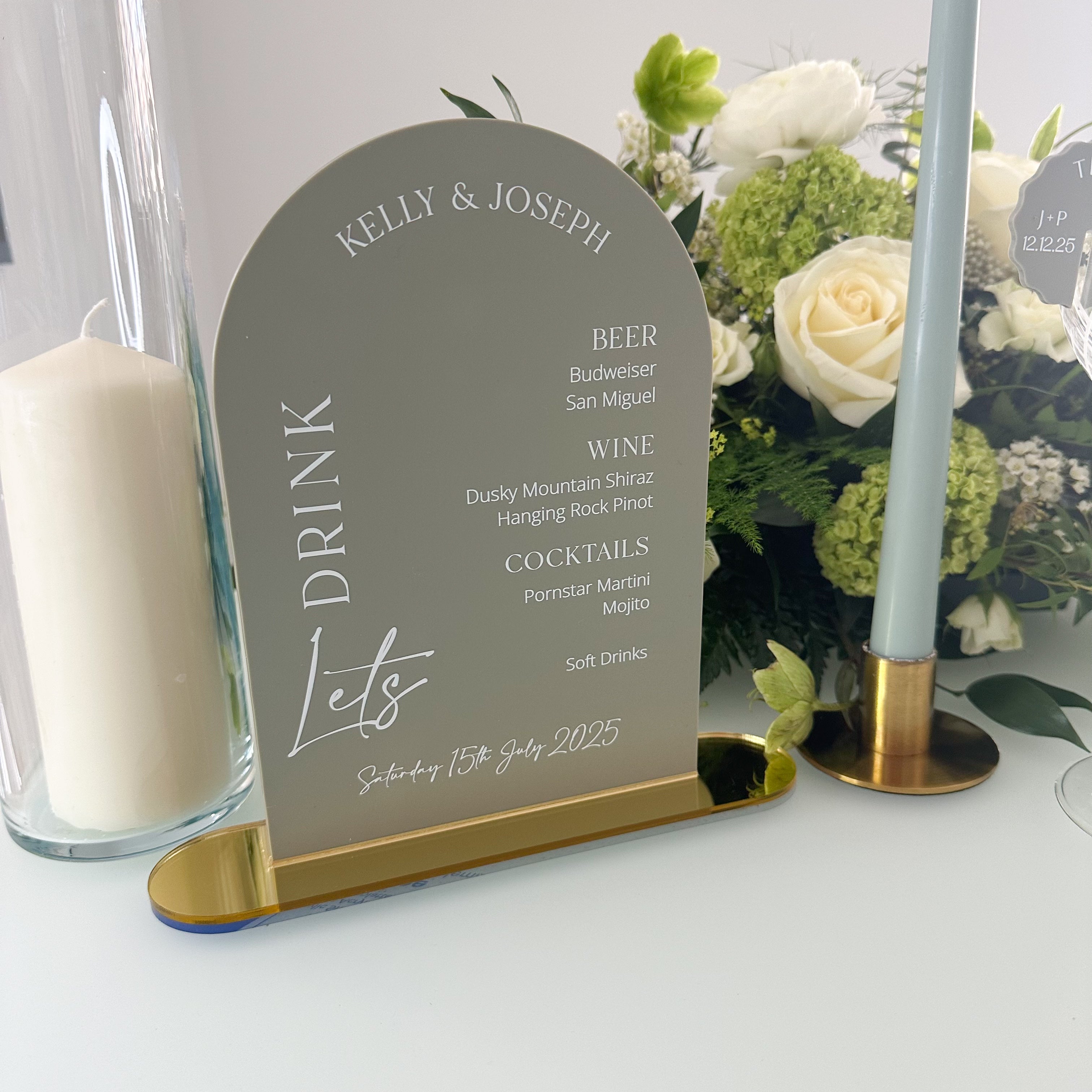 A gray sign displaying a drink menu, with a candle and flowers in the background.