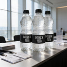 Three clear plastic water bottles with black labels are placed on a table, with a stack of business cards and a sign visible in the background.