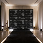 a black backdrop with white text and logos, situated in a room with a red carpet and a gold pole.