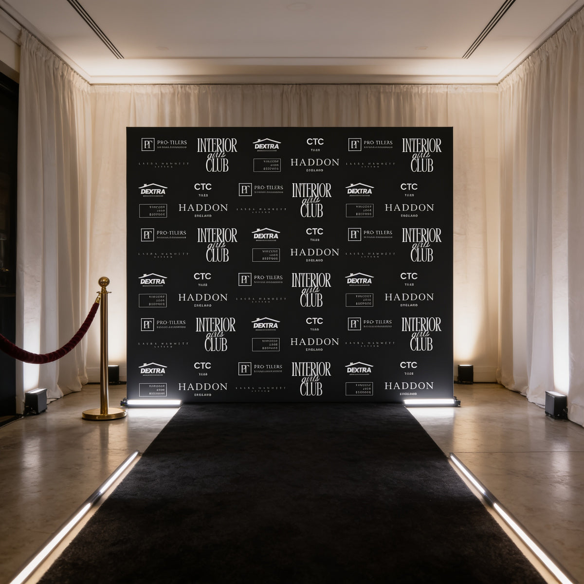 a black backdrop with white text and logos, situated in a room with a red carpet and a gold pole.