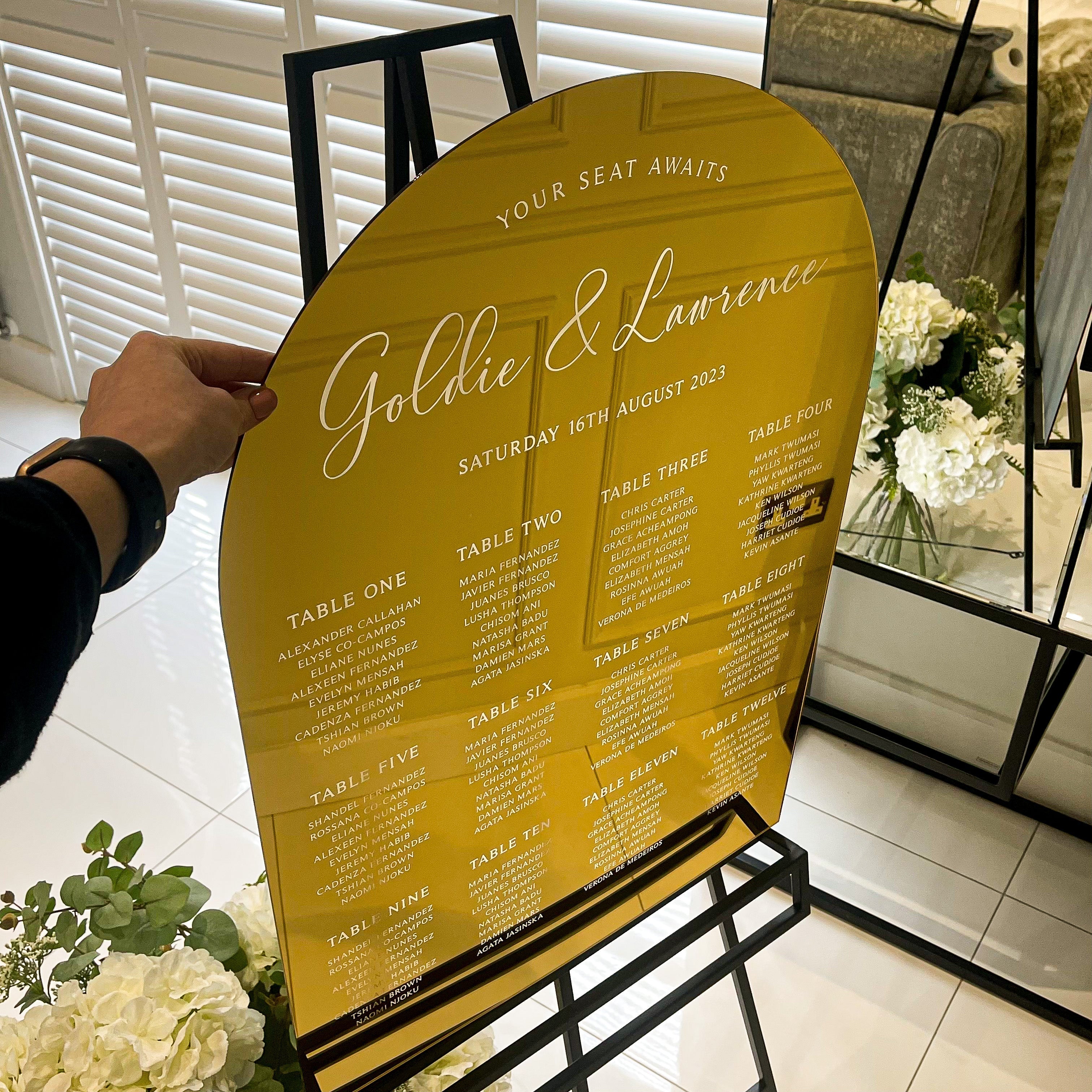 two large, gold-colored arch-shaped signs with text and a seating plan for a wedding reception.