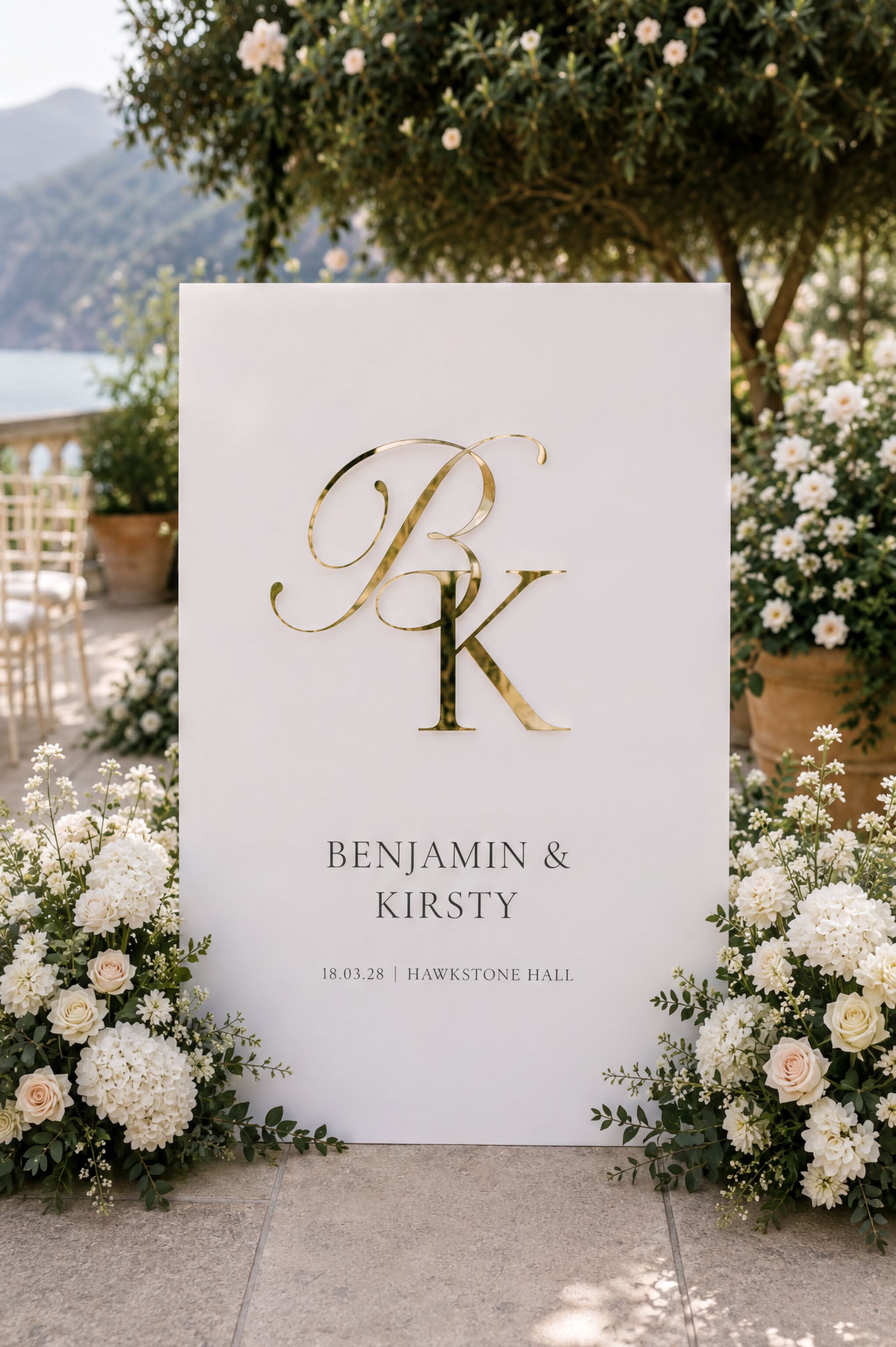 a white sign with a gold letter "P" and the names "BENJAMIN & KIRSTY" written on it, surrounded by white flowers and greenery.