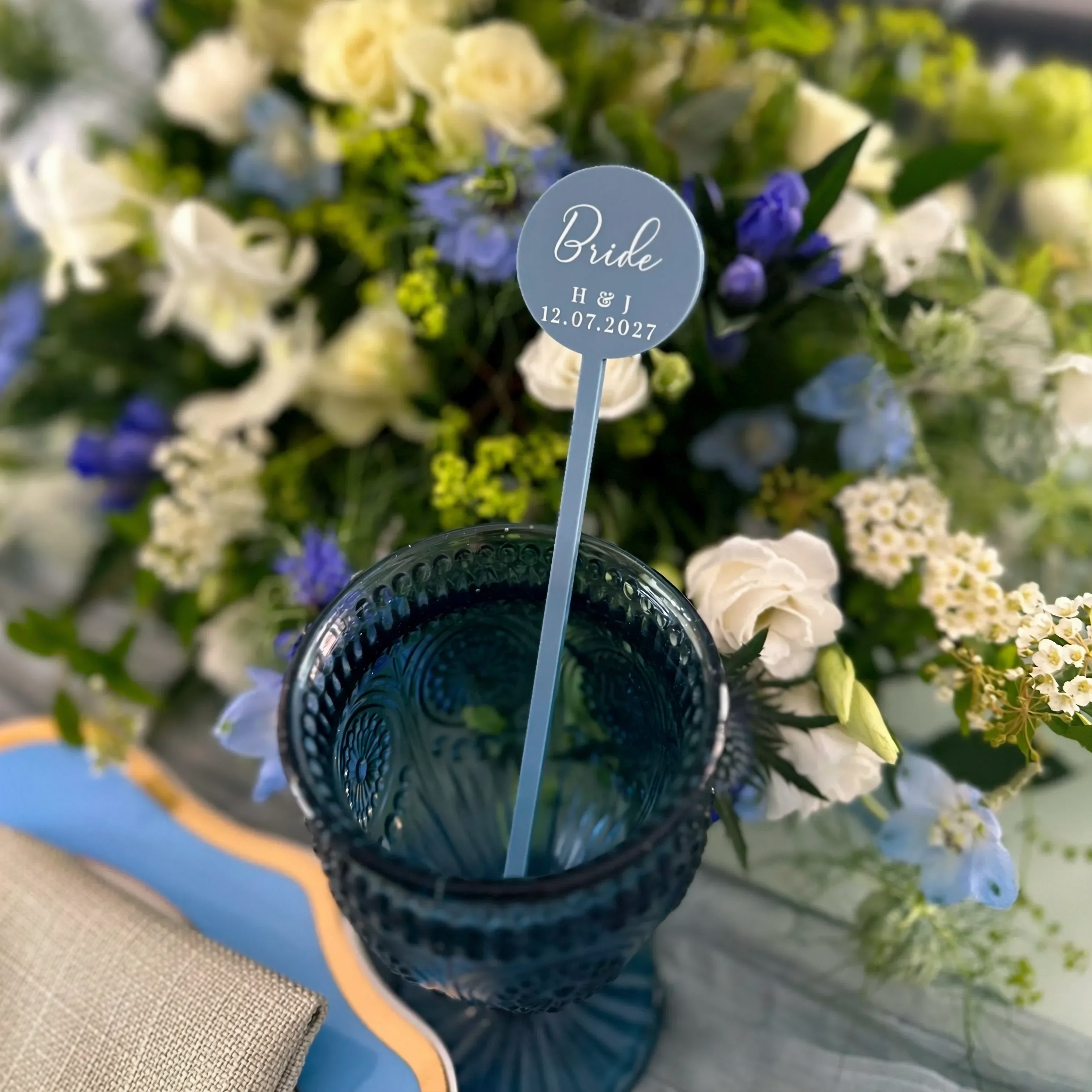 A blue glass with a blue straw is placed on a table with a floral arrangement in the background.