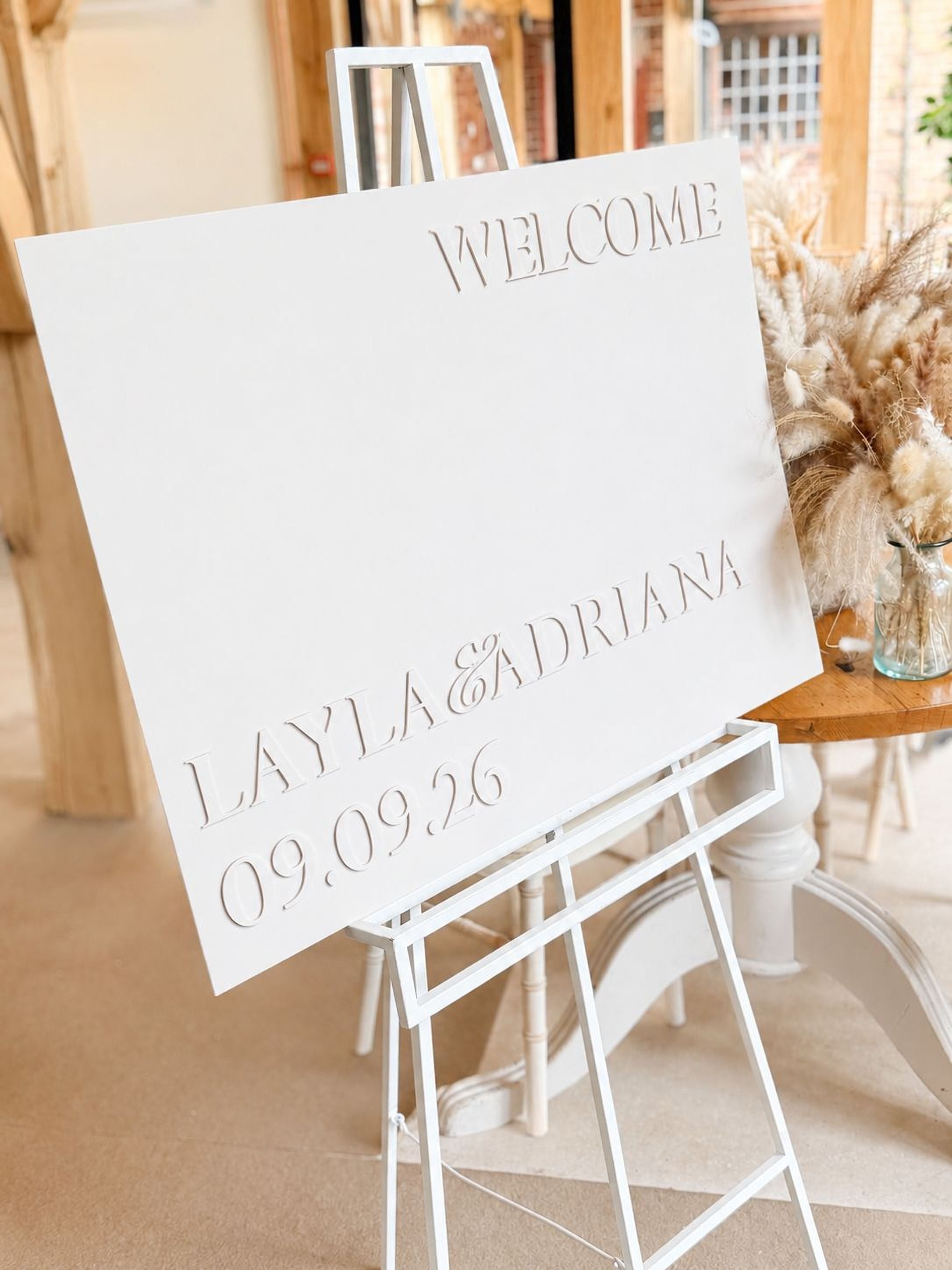 A white sign with the text "WELCOME LAYLA & ADRIANA 09.09.26" is displayed on a white easel.