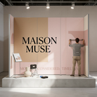 A man is standing in front of a large display with the word "MAISON MUSE" written on it, and there are some objects on the display, including a suitcase and a vase.
