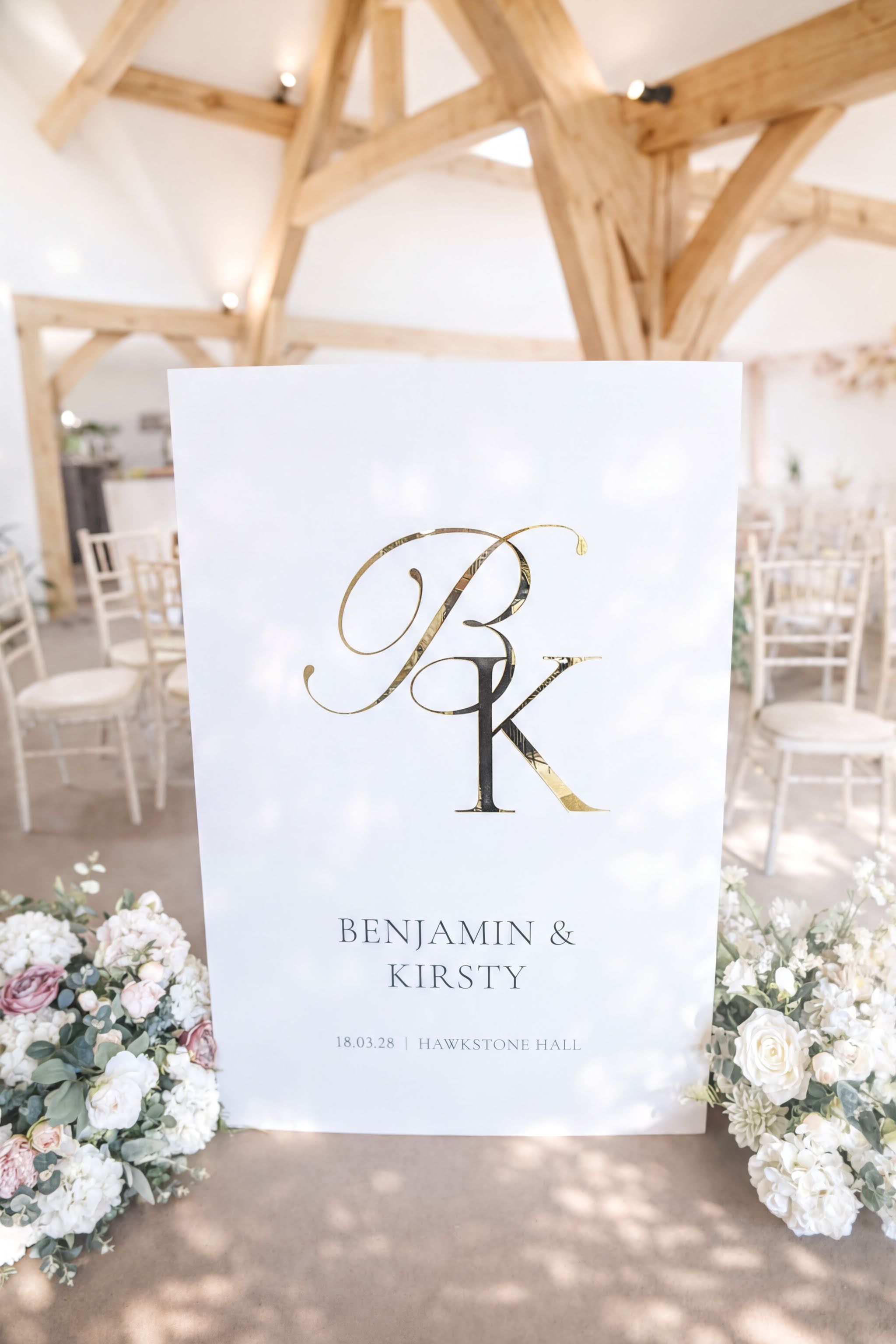 A white sign with the initials "BK" and the names "Benjamin & Kirsty" is displayed in a room with wooden beams and white chairs.