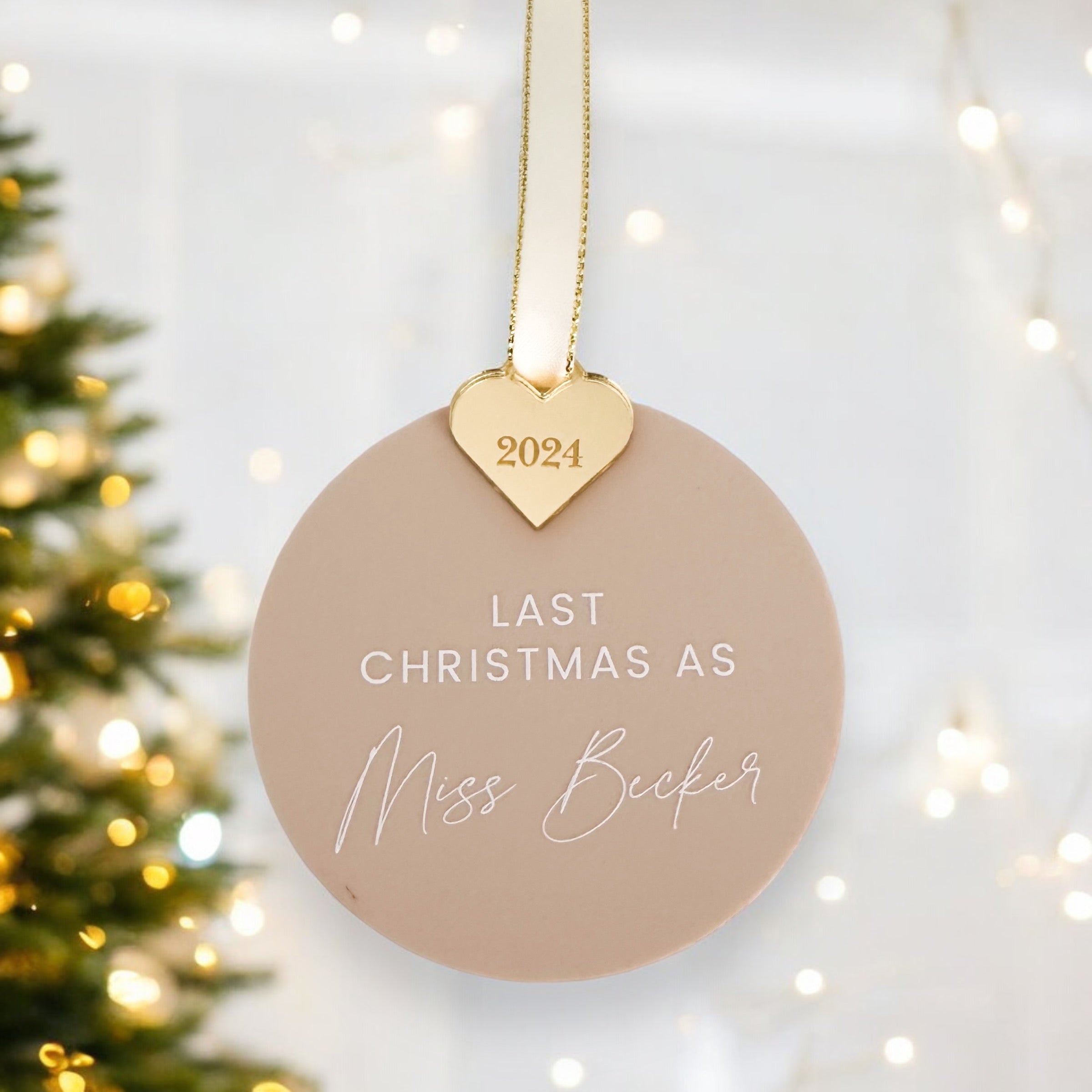 A white Christmas ornament with a gold heart-shaped ornament hanging from a string, displaying the personalised text "LAST CHRISTMAS AS Miss Smith" and the year "2024" on it.